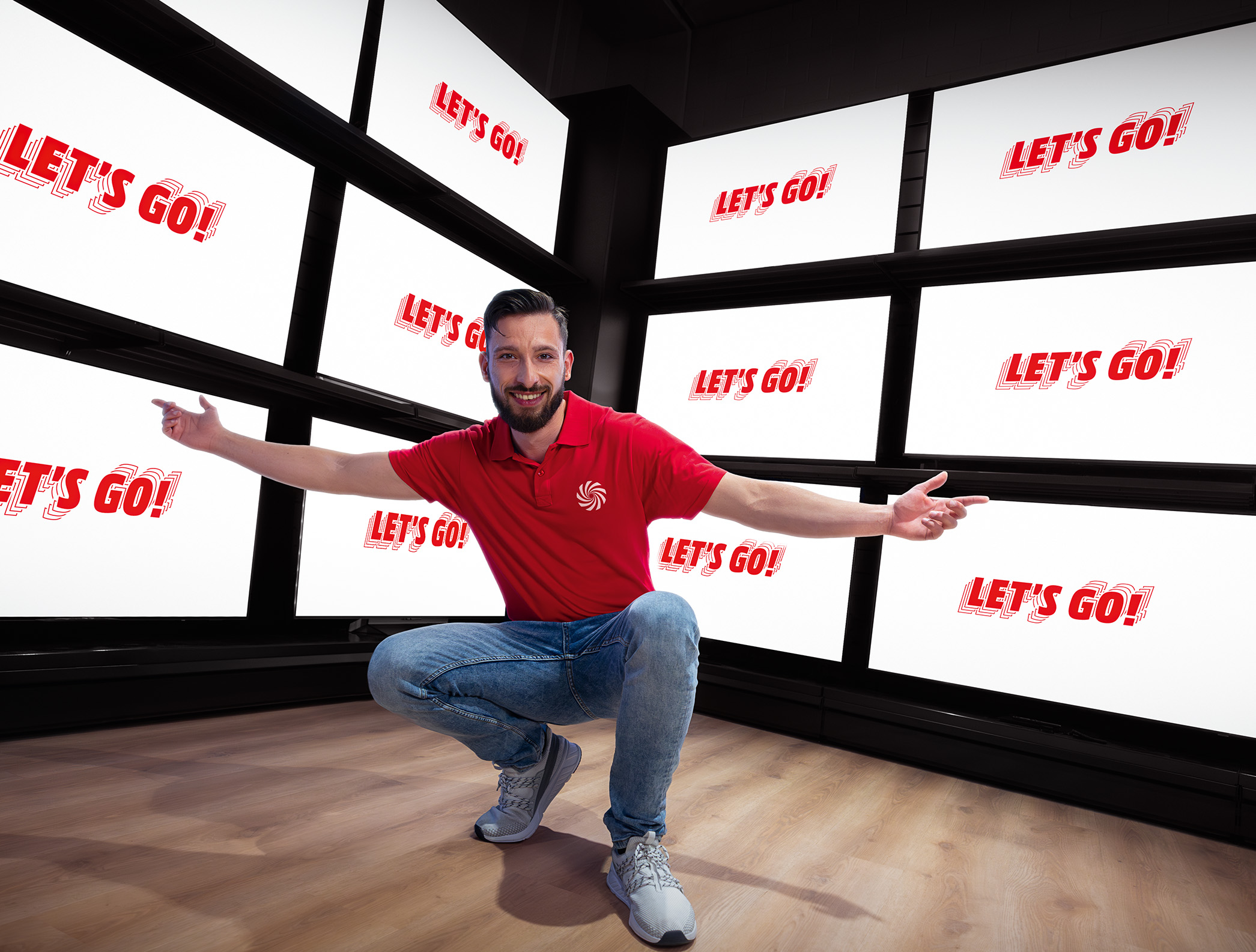 Verkäufer hockt im Markt vor einer TV-Wand mit Screens, worauf „Let’s Go“ steht.